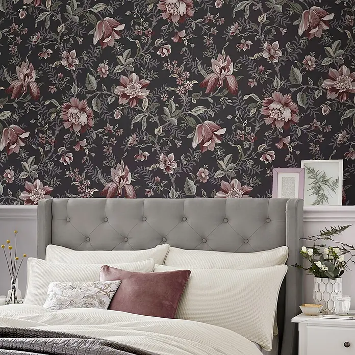 Laura Ashley Vliestapete Exotischer Garten - Grau, Floral, 1.000 x 52 cm - Dunkle Blumentapete mit roten und grünen Blumen hinter einem Bett mit grauem Kopfteil.