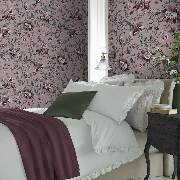 Laura Ashley Vliestapete Vliestapete Exotischer Garten - Rot, Floral, 10 x 0,52 m - Bett mit hellgrüner Rüschenbettwäsche, dunkelgrünem Kissen und burgunderroter Strickdecke im Schlafzimmer mit Blumentapete.