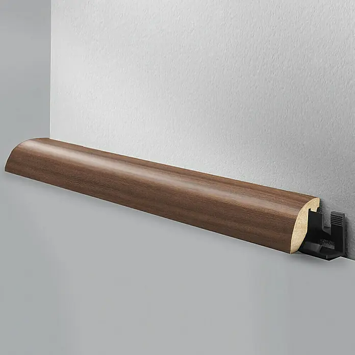 LOGOCLIC Viertelstab Nussbaum - 260 cm x 23 mm x 29 mm, MDF - Ein braunes, abgerundetes Sockelleiste-Profil ist an einer grauen Wand befestigt. Das Profil wird durch einen schwarzen Kunststoffhalter gehalten.