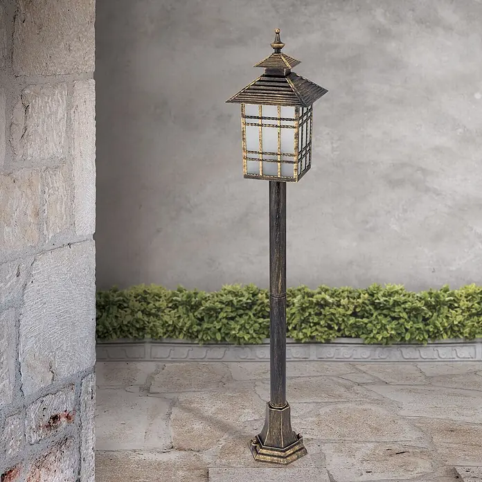 Orion Leuchten Außenlaterne Kiko - 60 W, Farbe Gehäuse: Bronze, Ohne Leuchtmittel - Eine verzierte, dunkelbronzefarbene Gartenlaterne mit quadratischem Leuchtenkopf und mattierten Glasscheiben steht auf einer gepflasterten Terrasse neben einer Steinmauer und Hecke.