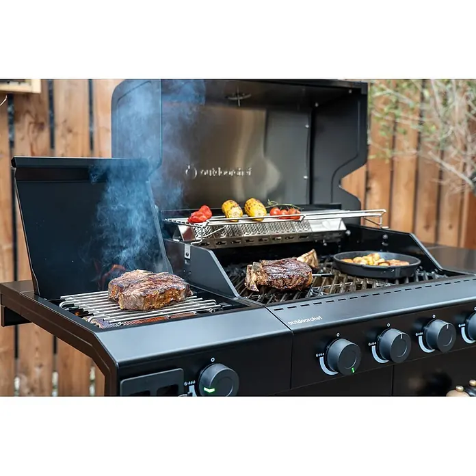 Outdoorchef Gasgrill Heat C435 - Grillfläche (B x T): 68 x 48 cm, 12,4 kW, Anzahl Brenner: 4 Stk. - Ein schwarzer Gasgrill mit Steaks, Mais, Paprika und Tomaten auf dem Rost, Rauch steigt auf. Kartoffeln sind in einer Pfanne auf dem Grill.