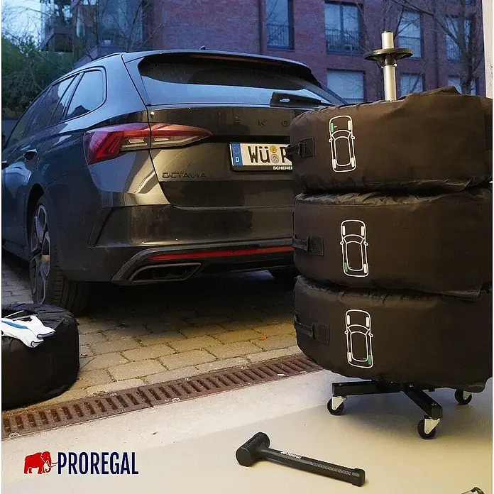 Proregal Reifenregal Fahrbarer Felgenbaum CARRERO - Stecken, Schwarz - Schwarze Autoreifen-Aufbewahrungstaschen mit Auto-Symbolen, auf einem mobilen Regal neben einem Fahrzeug, mit einem Gummihammer.