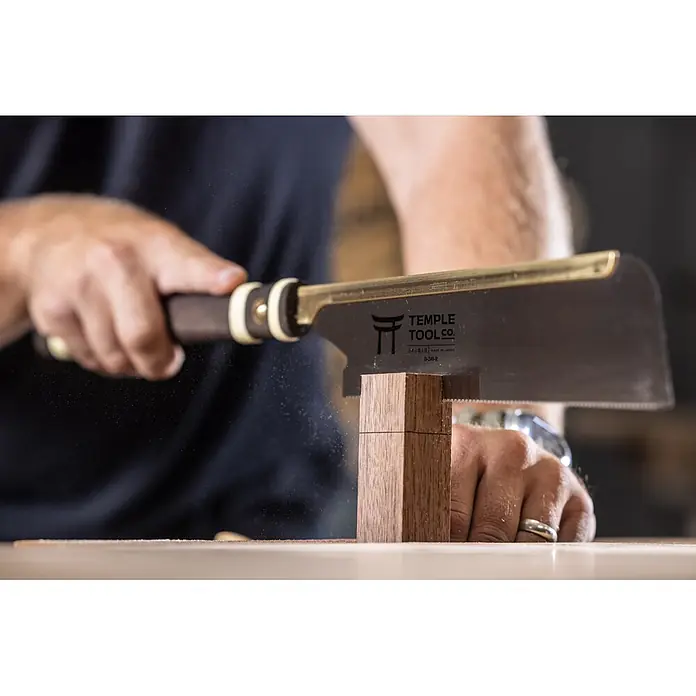Temple Tool Japansäge Dozuki Rip Cut - Blattlänge: 240 mm - Eine Person sägt mit einer japanischen Zugsäge mit dunklem Griff und Messingrücken ein Holzstück, wobei Sägespäne entstehen.