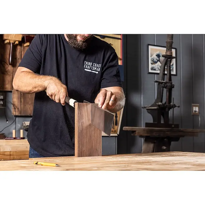 Temple Tool Japansäge Ryoba - Blattlänge: 240 mm - Handwerker sägt ein dunkles Holzbrett mit einer Handsäge, Sägespäne fliegen.