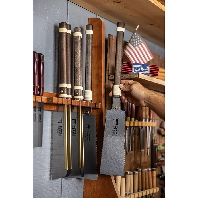 Temple Tool Japansäge Ryoba - Blattlänge: 240 mm - Eine Hand greift nach einer japanischen Zugsäge mit dunklem Holzgriff und hellem Band, die mit anderen Sägen an einem Regal in einer Werkstatt hängt.