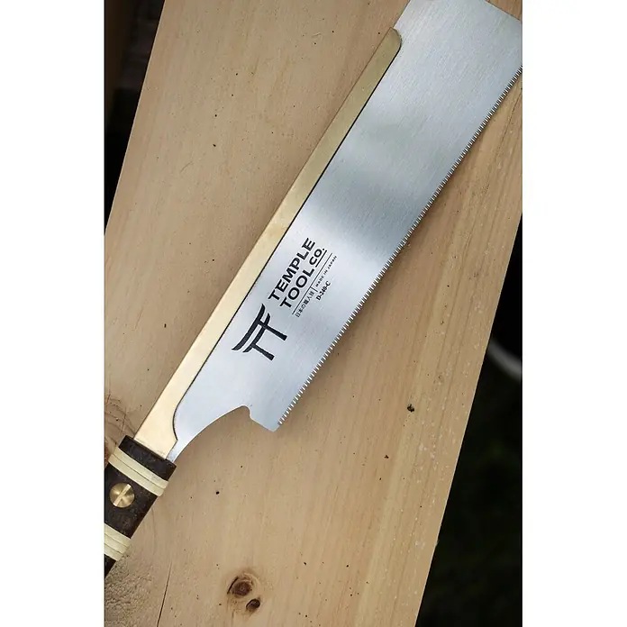 Temple Tool Japansäge Dozuki Cross Cut - Blattlänge: 240 mm - Eine japanische Zugsäge mit silberner Klinge, Messingrücken und dunklem Griff mit hellen Ringen, auf hellem Holz liegend.