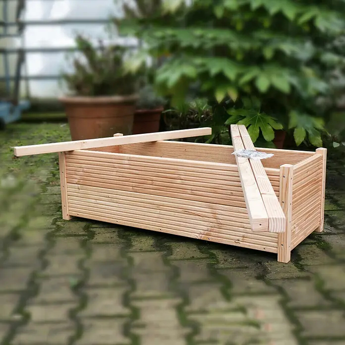 Helix Fertighecke Hecke am laufenden Meter® Balkon Komplettset - Hedera helix 'Woerneri', 100 x 20 x 166 cm - Unmontierter, heller Blumenkasten aus Holz auf Pflastersteinen mit Pflanzen im Hintergrund.