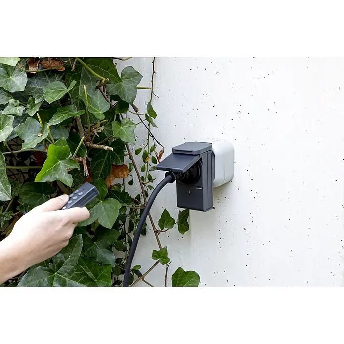 REV Funkschalter Set 2+1 außen 2 Stk. -  - Smarte Außensteckdose (dunkelgrau-weiß) an einer Wand, mit Kabel und Fernbedienung in der Hand.