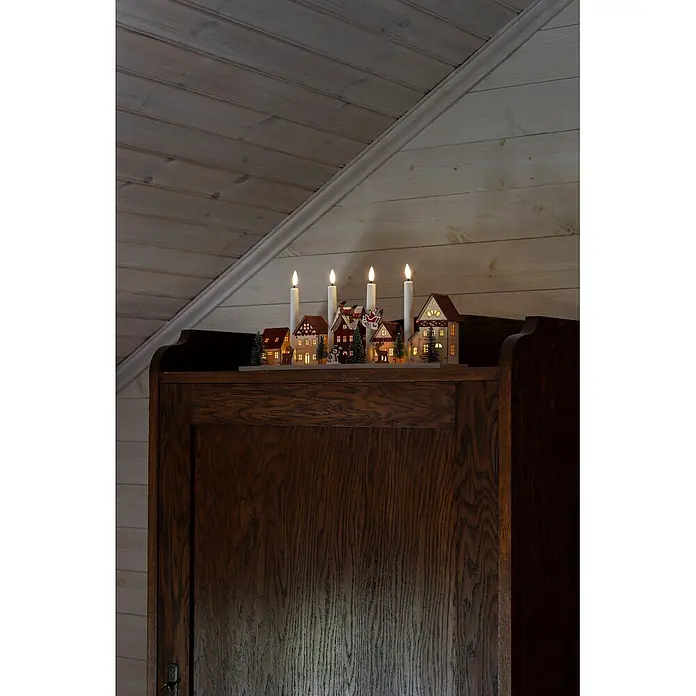 Konstsmide LED-Holzsilhouette Miniaturhäuser - 47 x 11 x 20,5 cm, Braun, Batteriebetrieben, Warmweiß - Beleuchtete Weihnachtsdorf-Dekoration mit vier Kerzenlichtern auf einem dunklen Holzschrank vor holzgetäfelter Wand.