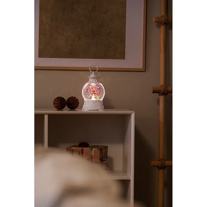 Konstsmide LED-Schneelaterne Schneemann mit Riesenrad - Innen, Weiß, 12,6 x 7,7 x 21 cm, Batteriebetrieben - Eine beleuchtete weiße Weihnachts-Laternen-Schneekugel mit bunten Figuren steht auf einem weißen Regal in einem festlichen Raum.