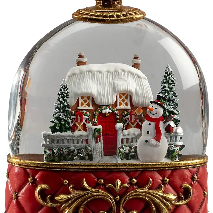 Konstsmide LED-Schneelaterne Schneemann mit Haus - Innen, Rot, 15,5 x 9 x 20 cm, Batteriebetrieben - Eine Weihnachtsschneekugel mit einer Winterdorfszene mit verschneitem Häuschen, Schneemann und Bäumen, auf einem roten gesteppten Sockel mit Goldakzenten.
