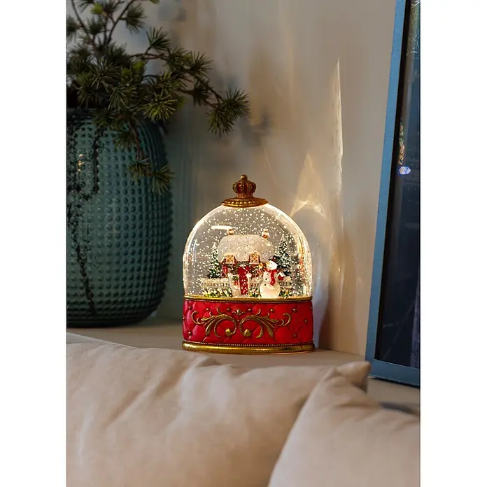 Konstsmide LED-Schneelaterne Schneemann mit Haus - Innen, Rot, 15,5 x 9 x 20 cm, Batteriebetrieben - Beleuchtete festliche Schneekugel mit rotem und goldenem Ziersockel, zeigt eine Winterlandschaft mit Haus und Schneemann auf einer Oberfläche.