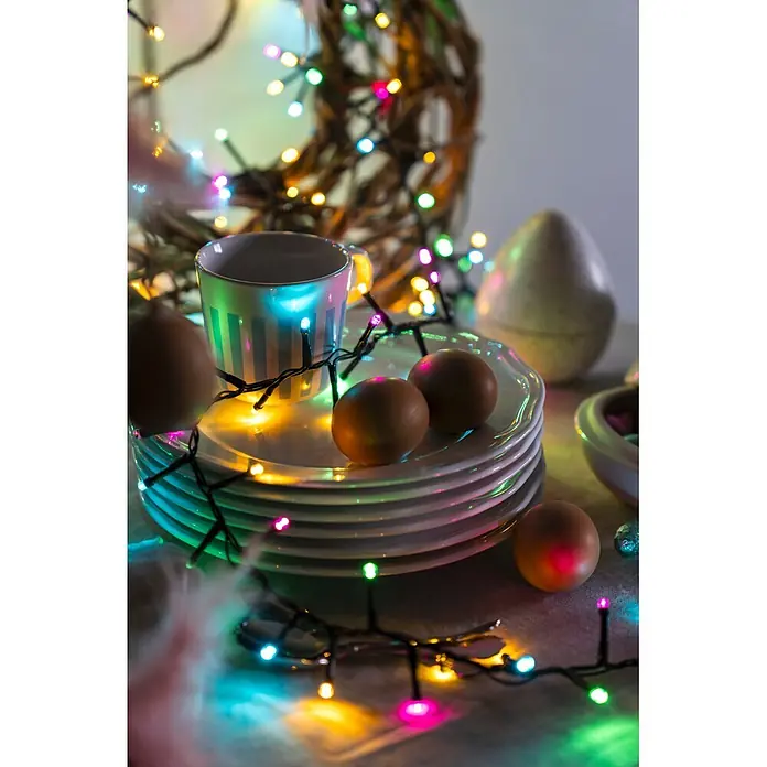 Konstsmide LED-Clusterlichterkette Pastell - Außen, Länge: 8,78 cm, 400 Stk., Netzbetrieben - Eine festliche Tischdekoration mit braunen Eiern auf weißen Tellern, einer gestreiften Tasse und bunten Lichterketten, die eine fröhliche Atmosphäre schaffen.