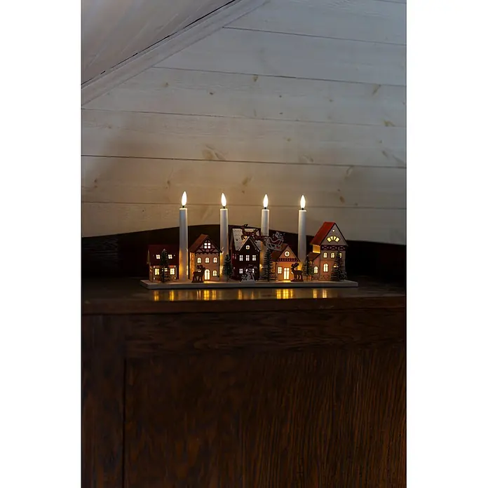 Konstsmide LED-Holzsilhouette Miniaturhäuser - 47 x 11 x 20,5 cm, Braun, Batteriebetrieben, Warmweiß - Beleuchtete Weihnachtslandschaft mit vier Kerzen auf dunklem Holz, vor einer hellen Holzwand.