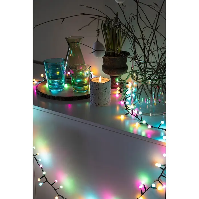 Konstsmide LED-Clusterlichterkette Globe Pastell - Außen, Länge: 4,38 m, 200 Stk., Netzbetrieben - Bunte Lichterkette, die über eine Oberfläche drapiert ist und dekorative Gläser, eine Kerze und Zweige beleuchtet.