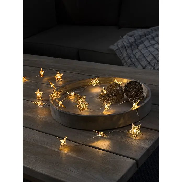 Konstsmide LED-Lichterkette - Innen, Lichtfarbe: Warmweiß, Anzahl Leuchtmittel: 20 Stk., Batteriebetrieben - Goldene sternförmige LED-Lichterkette spendet warmes Licht und schmückt einen Holztisch mit Tannenzapfen in gemütlicher Atmosphäre.