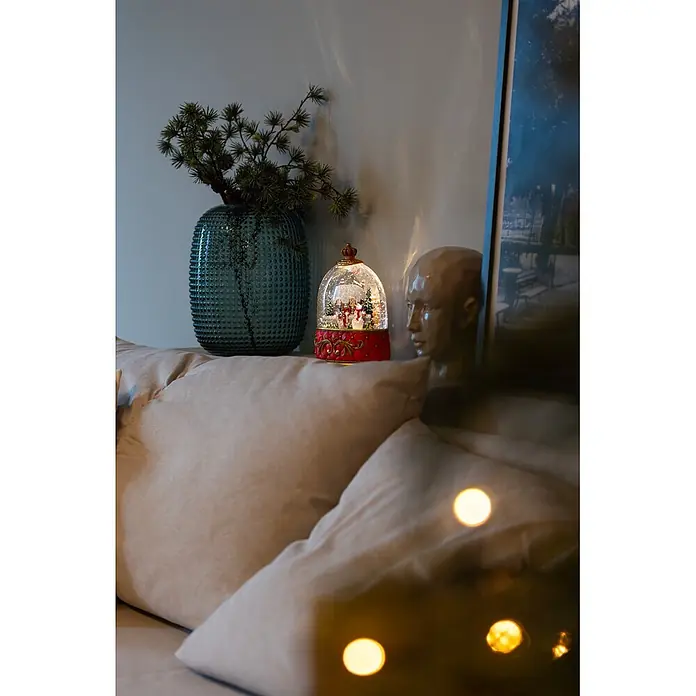 Konstsmide LED-Schneelaterne Schneemann mit Haus - Innen, Rot, 15,5 x 9 x 20 cm, Batteriebetrieben - Eine festliche, beleuchtete Schneekugel mit rotem Sockel und goldenen Akzenten, die eine Winterszene zeigt, auf Kissen platziert.