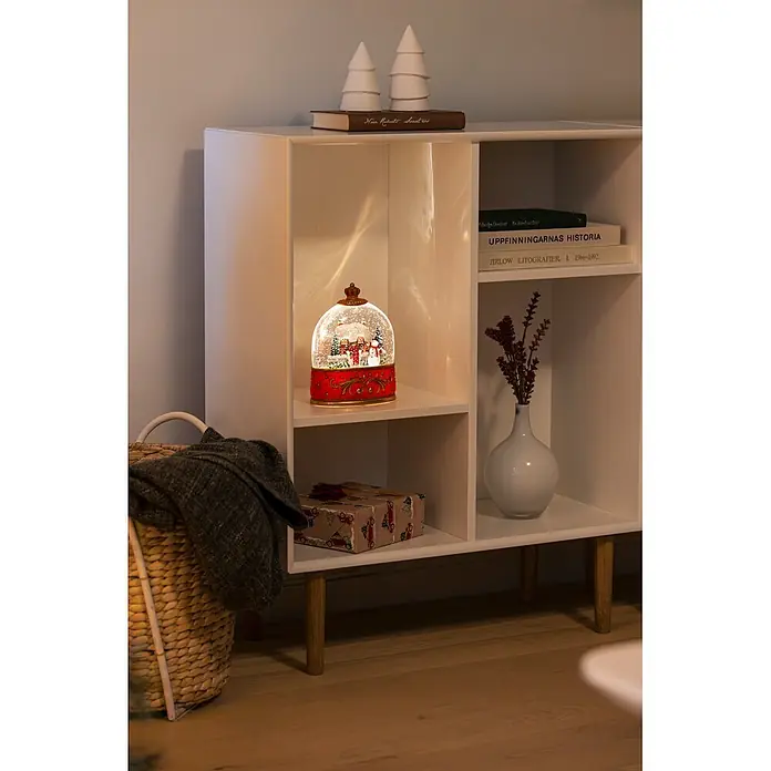 Konstsmide LED-Schneelaterne Schneemann mit Haus - Innen, Rot, 15,5 x 9 x 20 cm, Batteriebetrieben - Weißes Regal mit vier Fächern und hellen Holzbeinen, das eine beleuchtete Schneekugel und Bücher enthält.