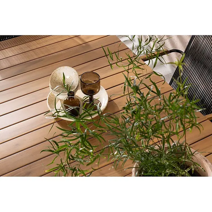 Ebuy24 Gartentisch-Set Holmbeck - Gesamtumfang: 7 -tlg., L x B x H: 90 x 200 x 75 cm, Ø x H: 90 x 75 cm, Stahl Akazie, Stapelbar - Nahaufnahme eines hölzernen Lamellentisches für draußen mit Dekoschalen, Glas, Flasche und Topfpflanze.