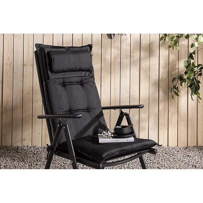 Ebuy24 Sitzauflage Cus - 1 Stk., L x B x H: 94 x 41 x 6 cm, Fasern, Schaum, Polyester., Schwarz - Schwarzer klappbarer Gartenliegestuhl mit weichen Polstern, einem Buch und einer Tasse, draußen auf Kies vor einer Holzwand.