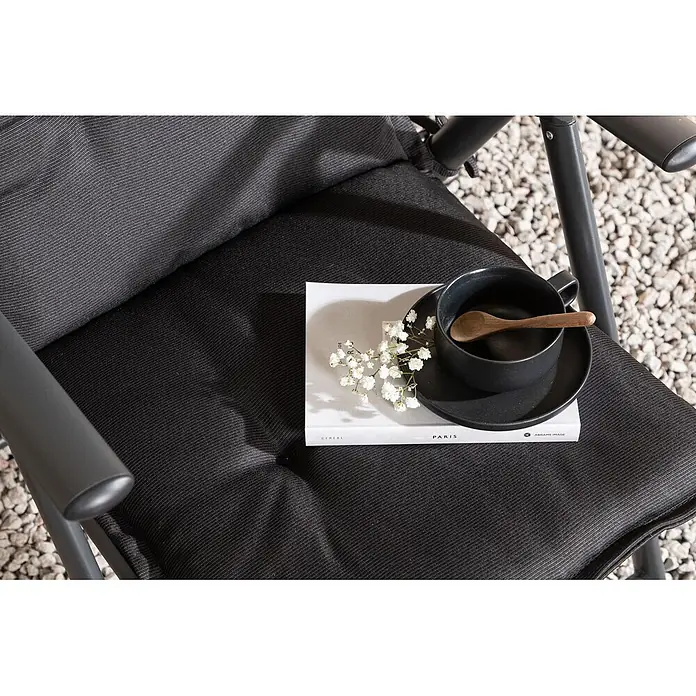 Ebuy24 Sitzauflage Cus - 1 Stk., L x B x H: 94 x 41 x 6 cm, Fasern, Schaum, Polyester., Schwarz - Schwarzer Outdoor-Klappstuhl mit bequemen Polstern. Inszeniert mit Buch, Tasse und Blumen für eine entspannende Atmosphäre.