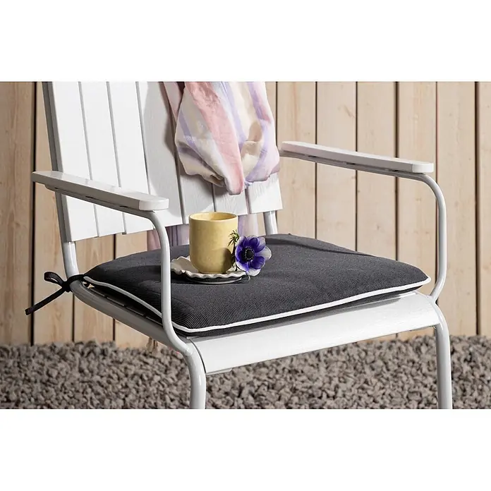Ebuy24 Sitzauflage Holmsund - 1 Stk., L x B x H: 47 x 41 x 4 cm, Fasern, Schaum, Polyester., Grau - Weißer Outdoor-Stuhl mit dunkelgrauem Kissen mit weißer Paspelierung, mit schwarzen Schleifen befestigt, draußen mit Tasse und Blume.