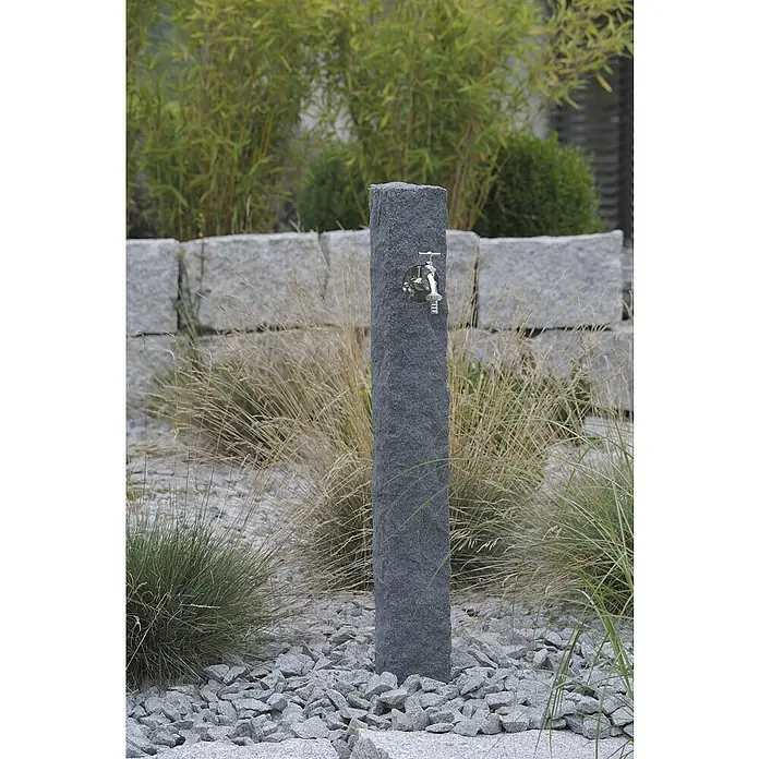 Arves Wasserzapfstelle Natura - Black Granitfarben, Höhe: 98 cm - Eine moderne Gartenszene mit einem stilvollen Wasserhahn aus dunklem Stein, umgeben von dekorativen Gräsern und Steinen.