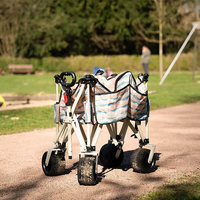 Home Deluxe Bollerwagen JOLA - L x B x H: 55 x 99 x 57 cm - Klappbarer Bollerwagen mit pfirsich-blau gestreiftem Stoff, schwarzem Griff und schmutzigen Rädern, auf einem Schotterweg im Freien.