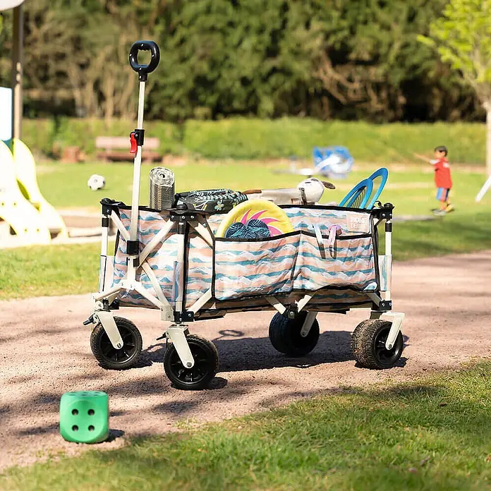 Home Deluxe Bollerwagen JOLA - L x B x H: 55 x 99 x 57 cm - Klappbarer Bollerwagen mit beigem Gestell und blau/orange gewelltem Stoff, gefüllt mit Spielzeug, im Park.