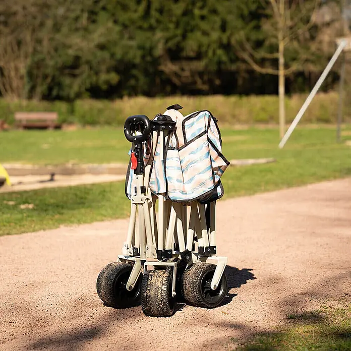 Home Deluxe Bollerwagen JOLA - L x B x H: 55 x 99 x 57 cm - Ein zusammengeklappter beiger Bollerwagen mit gemustertem Stoff und schwarzen Rädern steht auf einem Kiesweg im Freien.