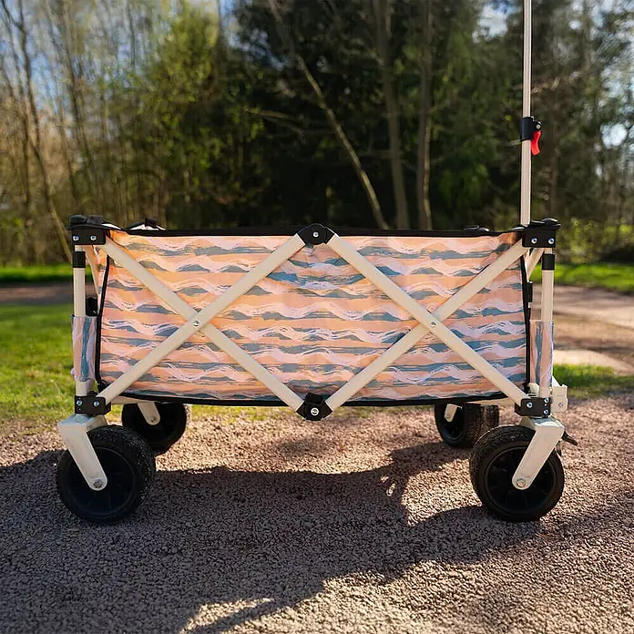 Home Deluxe Bollerwagen JOLA - L x B x H: 55 x 99 x 57 cm - Klappbarer Bollerwagen mit orange-blau gewelltem Stoffmuster und hellem Rahmen, auf einem Schotterweg im Freien.