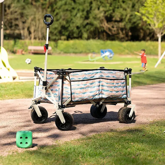 Home Deluxe Bollerwagen JOLA - L x B x H: 55 x 99 x 57 cm - Klappbarer Bollerwagen mit blau-orangem Wellenmuster-Stoff und hellem Gestell in einem Park.