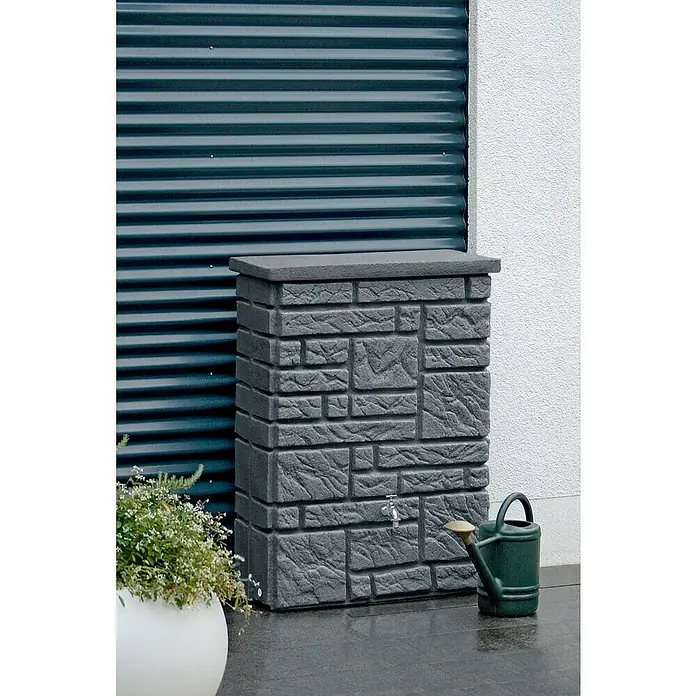 Arves Regenspeicher Maurano - 300 l, Black Granitfarben, 1 Stk. - Eine graue Regentonne in Steinoptik mit einem Wasserhahn, neben einer grünen Gießkanne und einem Topf mit Pflanzen, vor einem Rolltor.