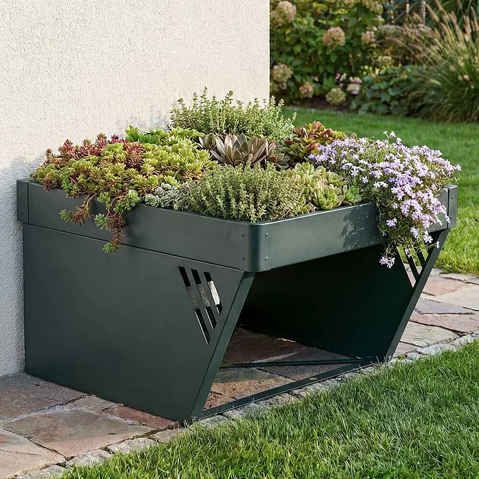 Proregal Mähroboter-Garage Mit Pflanzkasten - L x B x H: 70 x 60 x 40 cm, Grün, Ausführungseigenschaft: mit Pultdach, Gardena, WORX, Husqvarna, Einhell, Landxcape, Juka - Dunkelgrünes Metallbeet mit schrägem Design, gefüllt mit Sukkulenten und lila Blüten, an einer weißen Wand im Garten.
