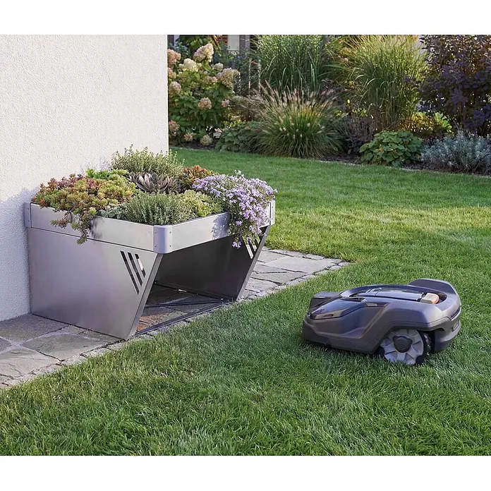 Proregal Mähroboter-Garage Mit Pflanzkasten - L x B x H: 70 x 60 x 40 cm, Silber, Ausführungseigenschaft: mit Pultdach, Gardena, WORX, Husqvarna, Einhell, Landxcape, Juka - Silberne Metallgarage für Mähroboter mit bepflanztem Gründach, neben einer weißen Wand. Ein dunkelgrauer Mähroboter steht auf dem Rasen.