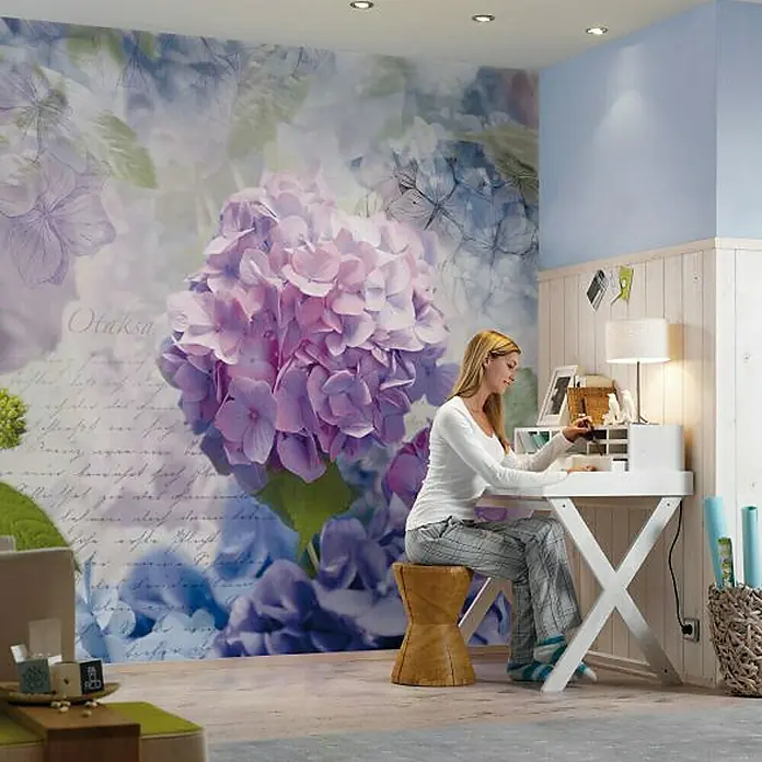 Komar Imagine Edition 3 - Stories Fototapete Otaksa - 8 -tlg., B x H: 368 x 254 cm, Papier - Frau am weißen Schreibtisch mit Blumenwandbild. Der Schreibtisch bietet Stauraum und ist mit einer Lampe und Dekorationen ausgestattet.
