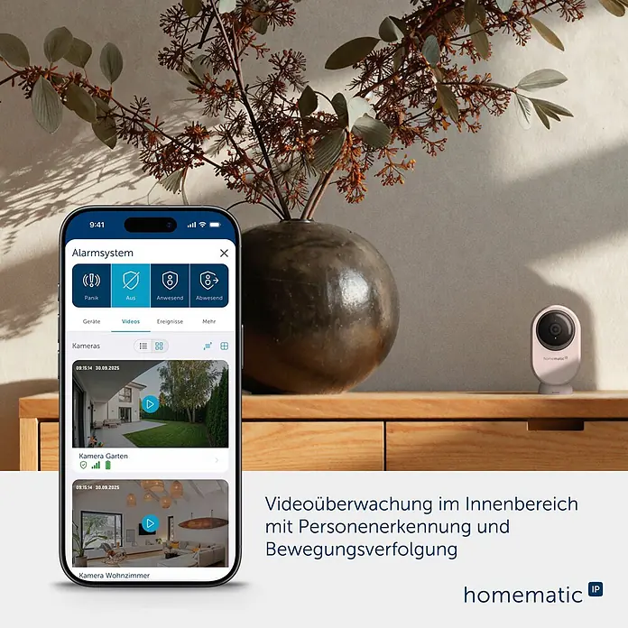 Homematic IP Innenkamera Smart Home HmIP-CI-PL - Weiß - Weiße Smart-Home-Kamera auf Holzfläche neben Smartphone mit Sicherheits-App und Videofeeds. Mit Personenerkennung & Bewegungsverfolgung.