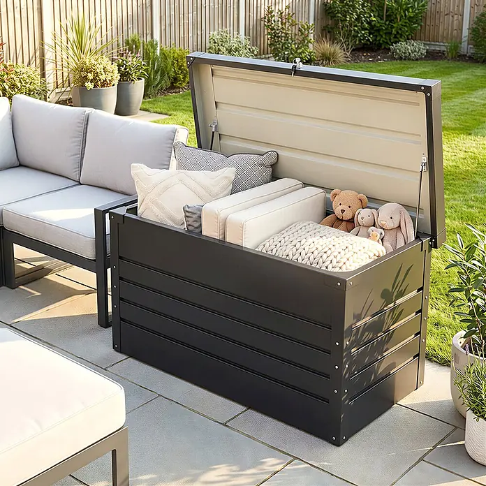 Outsunny Garten-Aufbewahrungsbox - Grau, Stahl - Dunkelgraue Outdoor-Aufbewahrungsbox, offen, gefüllt mit Kissen, Decke und Plüschtieren, auf einer Terrasse neben einem Sofa.