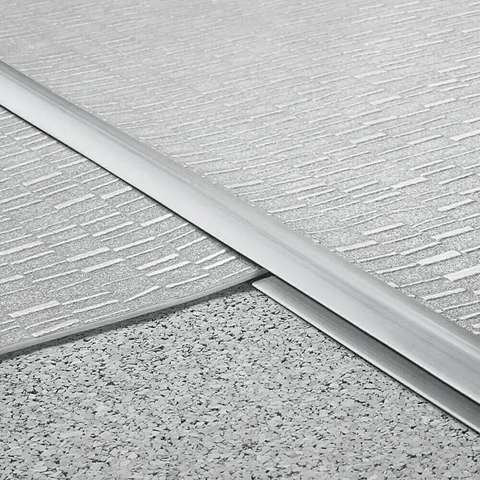 LOGOCLIC Doppeleinfassleiste - Grau, 1 m x 24 mm x 6 mm, Montageart: Kleben - Nahaufnahme einer Übergangsleiste zwischen zwei verschiedenen Bodenbelägen. Die Leiste ist aus Aluminium und trennt strukturierten von körnigem Boden.