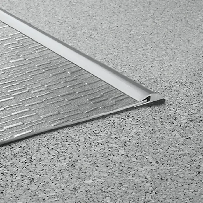 LOGOCLIC Einfassleiste - Grau, 1 m x 26 mm x 3 mm, Montageart: Kleben - Nahaufnahme eines Übergangsprofils zwischen zwei verschiedenen Bodenbelägen. Das Profil ist silberfarben und verbindet einen Teppich mit einem Steinboden.
