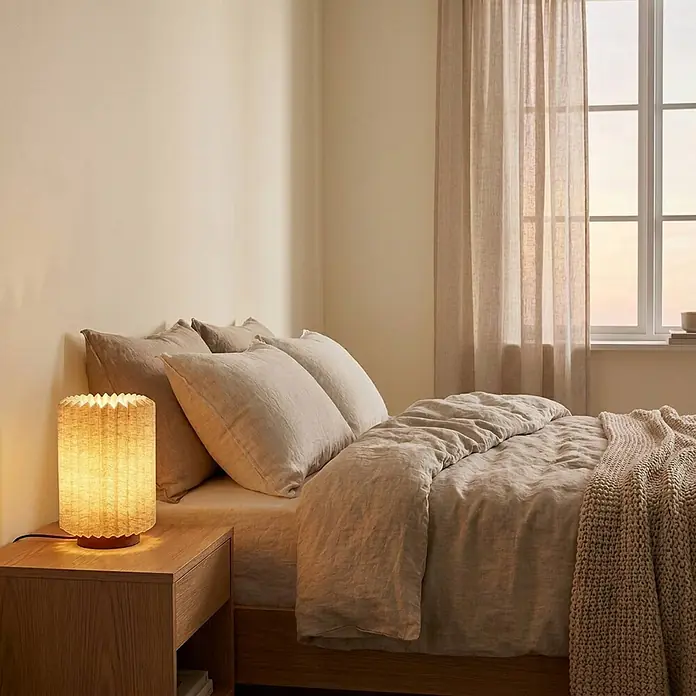 Lindby Tischleuchte Maveo - Holz dunkel, alu / grau / zink, E27 - Ein Bett mit beiger Leinenbettwäsche und einer gestrickten Decke, daneben ein Nachttisch aus Holz mit einer leuchtenden Faltlampe.