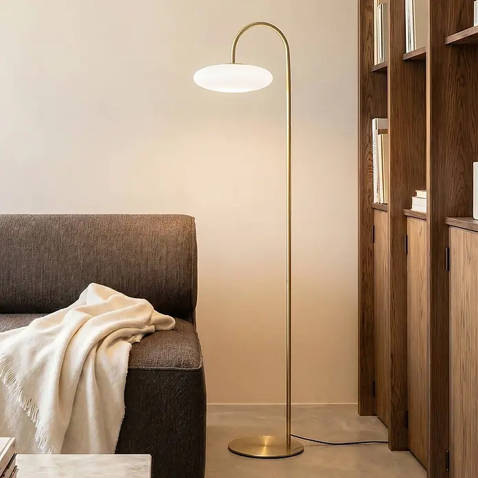 Lucande Stehleuchte Tenebra - messing / gold, weiß / opal - Goldfarbene Stehlampe mit gebogenem Stiel und flachem weißem Diffusor, beleuchtet ein Wohnzimmer neben einem dunklen Sofa.