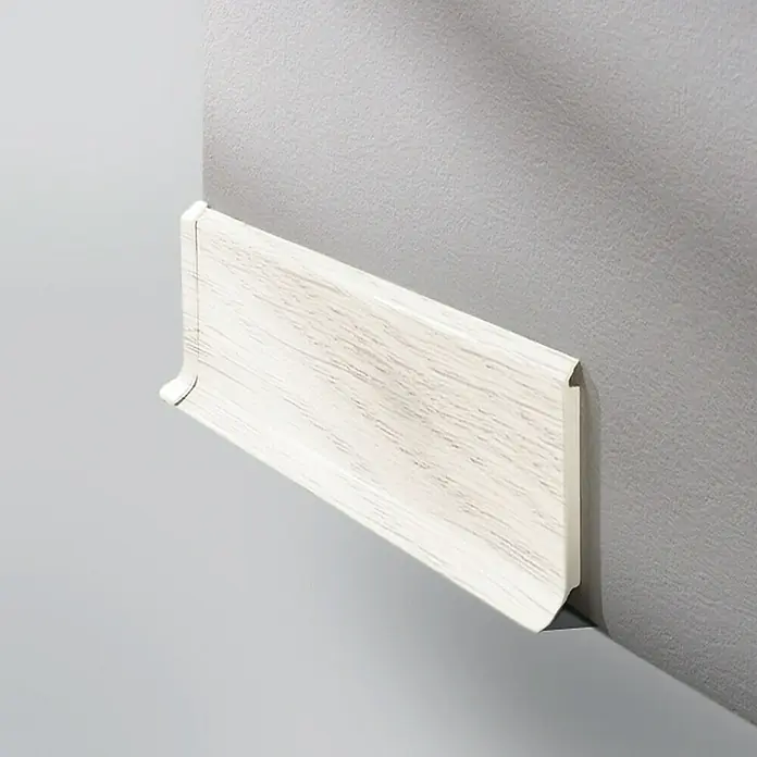 LOGOCLIC Sockelleiste HSL - 250 cm x 14 mm x 60 mm, Hartschaum, Esche Weiß - Eine weiße Holzfußleiste ist an einer grauen Wand befestigt. Die Leiste hat ein abgerundetes Profil und eine feine Holzmaserung.