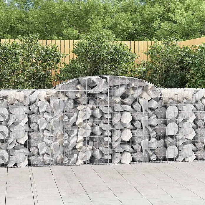 Furnicato Gabione - 50 x 300 x 120 cm, Metall, Maschenbreite: 50 mm, Maschenhöhe: 100 mm - Gabionenwand aus hellgrauen Steinen in Metallgeflecht, in einem Garten mit Holzzaun und grünen Pflanzen.