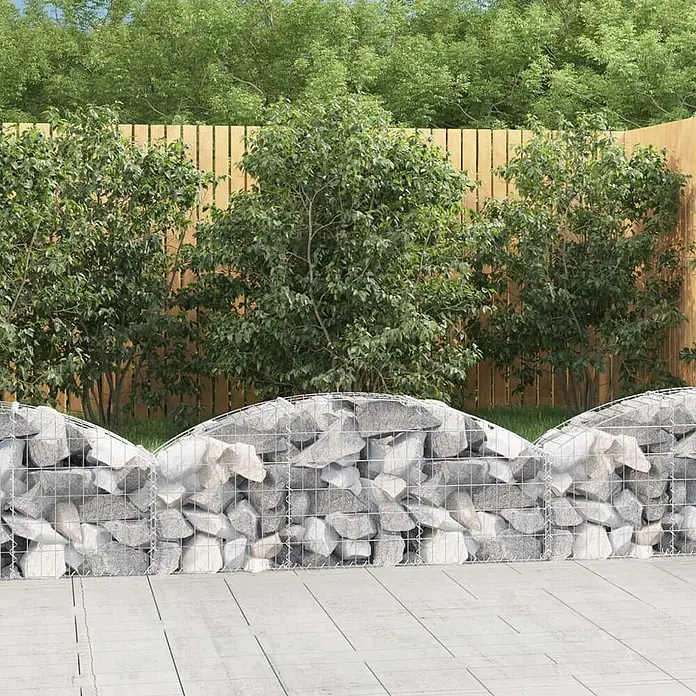 Furnicato Gabione - 30 x 150 x 60 cm, Metall, Maschenbreite: 50 mm, Maschenhöhe: 100 mm - Geschwungene Gabionenwand aus grau-weißen Steinen als dekorative Gartentrennung vor einem Holzzaun.