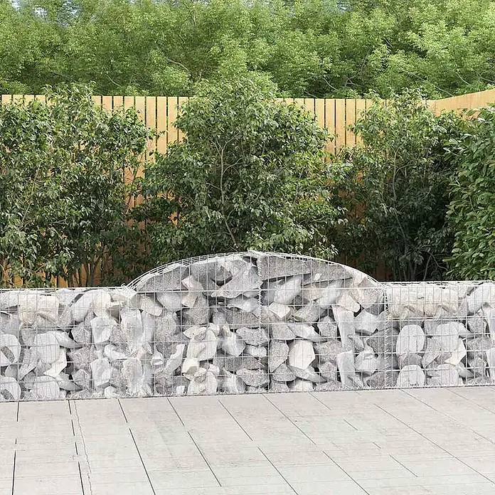 Furnicato Gabione - 30 x 300 x 80 cm, Metall, Maschenbreite: 50 mm, Maschenhöhe: 100 mm - Gabionenwand mit hellgrauen Steinen und gewölbter Oberseite, in einem Garten mit Holzzaun und grünen Bäumen.