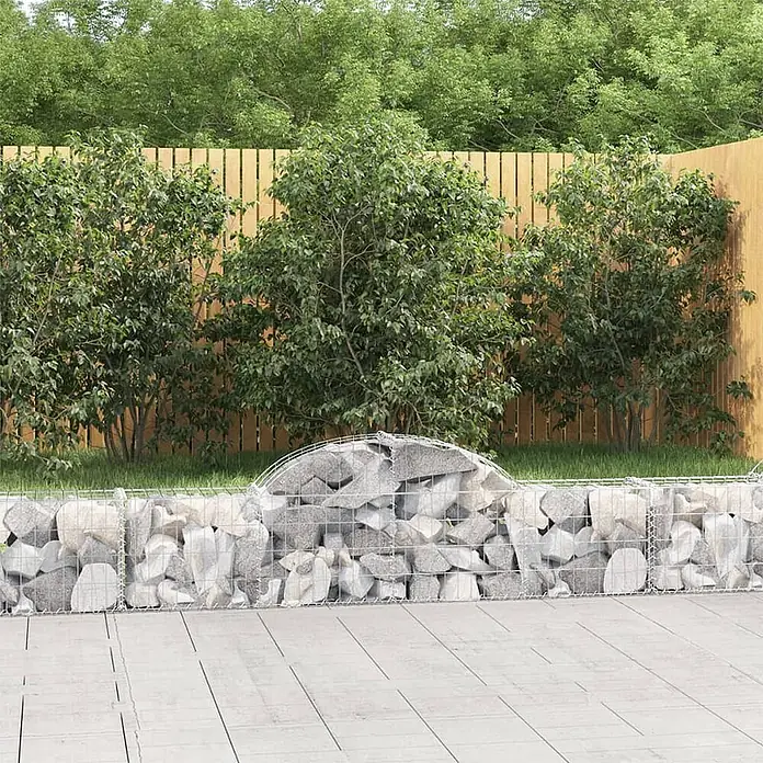 Furnicato Gabione - 30 x 200 x 60 cm, Metall, Maschenbreite: 50 mm, Maschenhöhe: 100 mm - Eine Gabionenwand, gefüllt mit grauen Steinen und mit einem gewölbten Oberteil, trennt einen gepflasterten Bereich von Gartenpflanzen und einem Holzzaun.