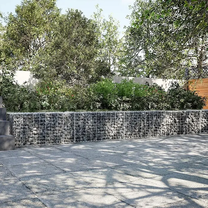 Furnicato Gabione - 800 x 50 x 50 cm, Metall, Maschenbreite: 50 mm, Maschenhöhe: 100 mm - Gabionen-Hochbeet mit Metallgitter, gefüllt mit grauen Steinen, trennt eine gepflasterte Terrasse von einem Garten mit Bäumen und Sträuchern.