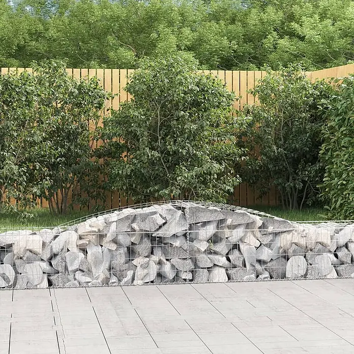 Furnicato Gabione - 60 x 400 x 50 cm, Metall, Maschenbreite: 50 mm, Maschenhöhe: 100 mm - Eine niedrige, wellenförmige Gabionenwand, gefüllt mit grauen Steinen, trennt eine graue Terrasse von einem Garten mit Holzzaun.