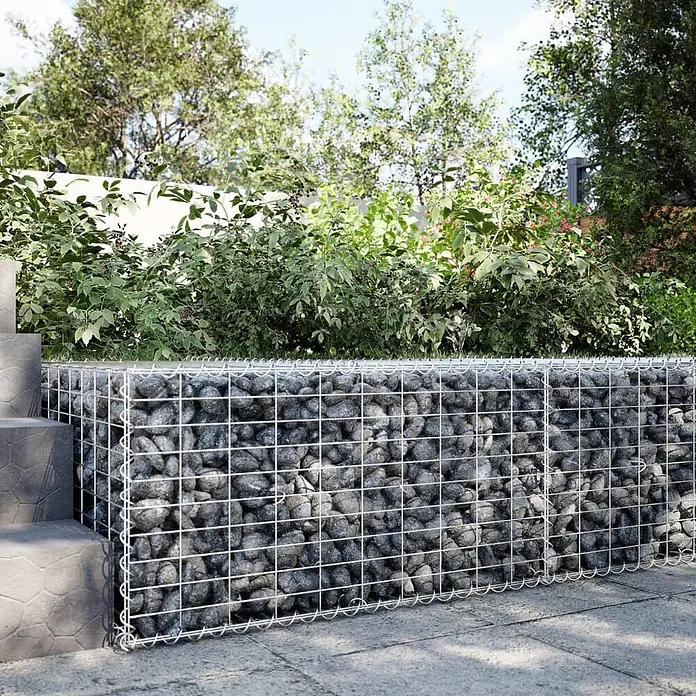 Furnicato Gabione - 100 x 100 x 50 cm, Metall, Maschenbreite: 50 mm, Maschenhöhe: 100 mm - Eine Gabionen-Stützmauer aus silbernem Drahtgeflecht, gefüllt mit grauen Steinen, in einem Garten neben Stufen und Pflaster.